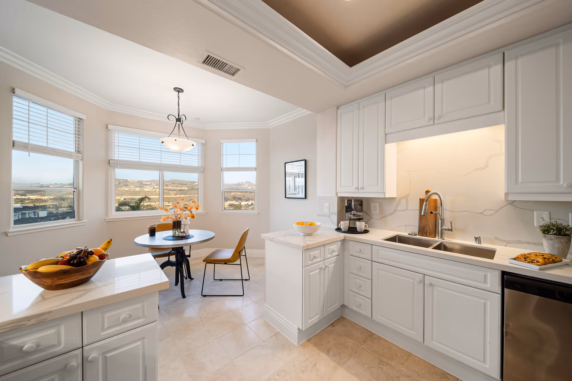 Bright kitchen area with white cabinetry, a sink, and a countertop with a bowl of lemons and a plate of cookies. Adjacent to the kitchen is a dining nook with a round table, two chairs, a vase with orange flowers, and large windows showing a scenic view of hills and buildings outside.