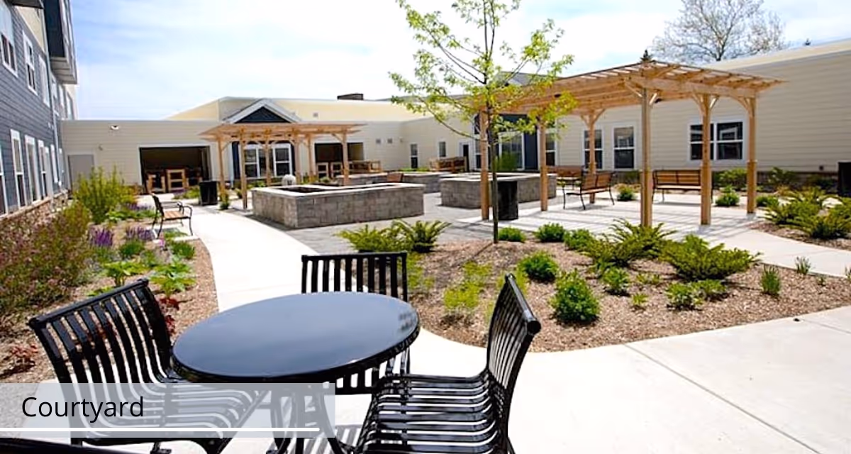 Outdoor courtyard with a round metal table and chairs, pergolas, raised planters and walkways at a senior living facility.