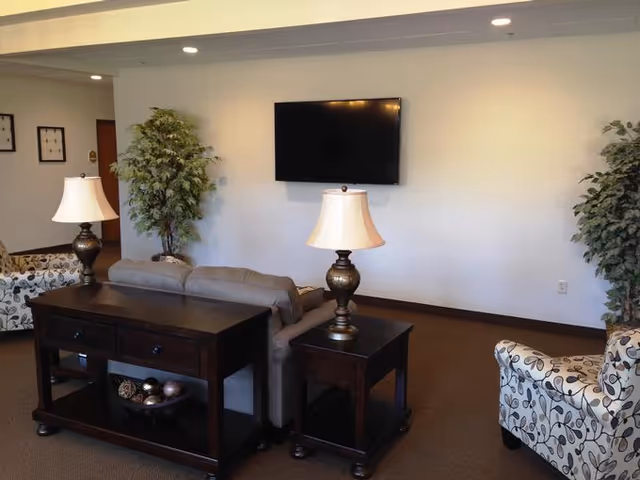 A cozy living room area with a beige sofa, two patterned armchairs, two table lamps with beige shades on dark wooden tables, a flat-screen TV mounted on a plain white wall, and two large potted plants on either side of the TV.