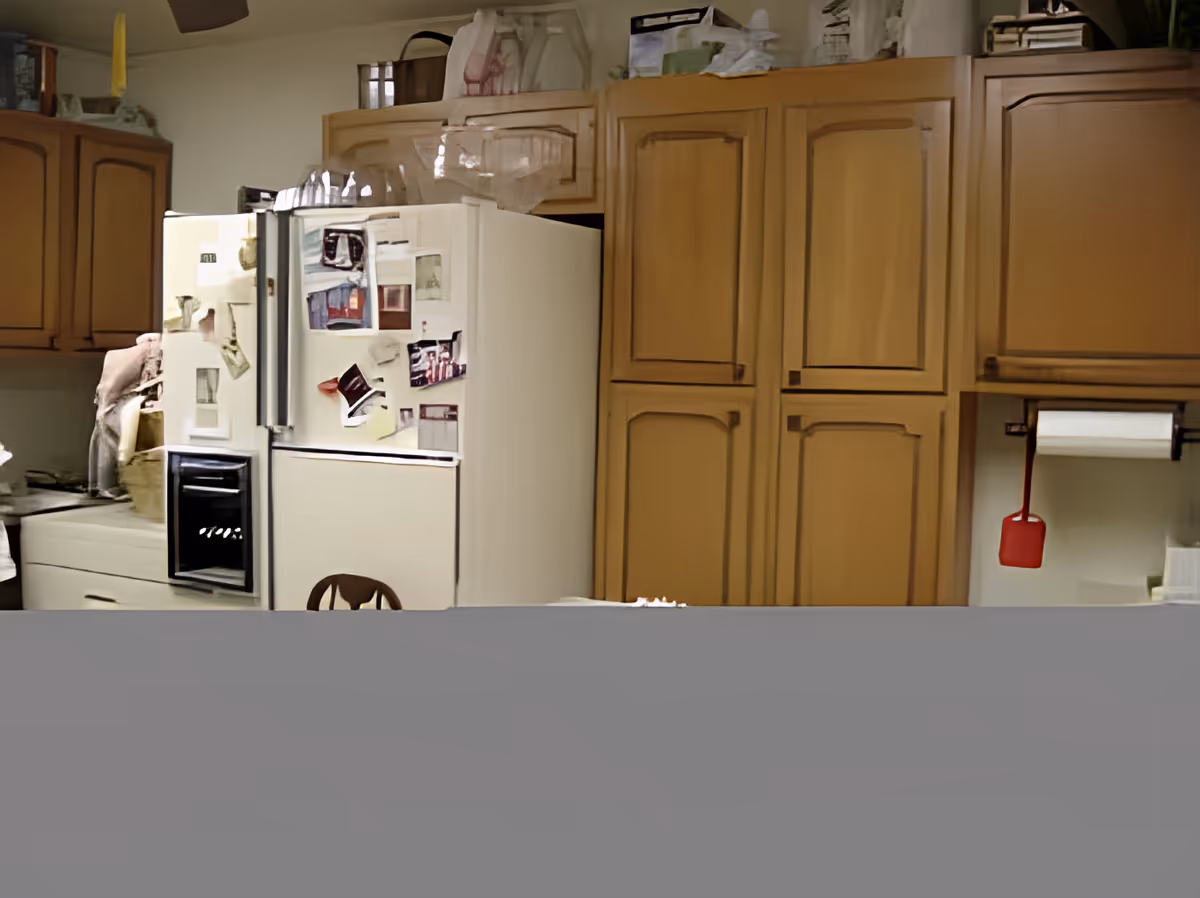 Interior view of a kitchen featuring wooden cabinets, a white refrigerator covered with various photos and papers, and a paper towel holder with a red spatula hanging from it.