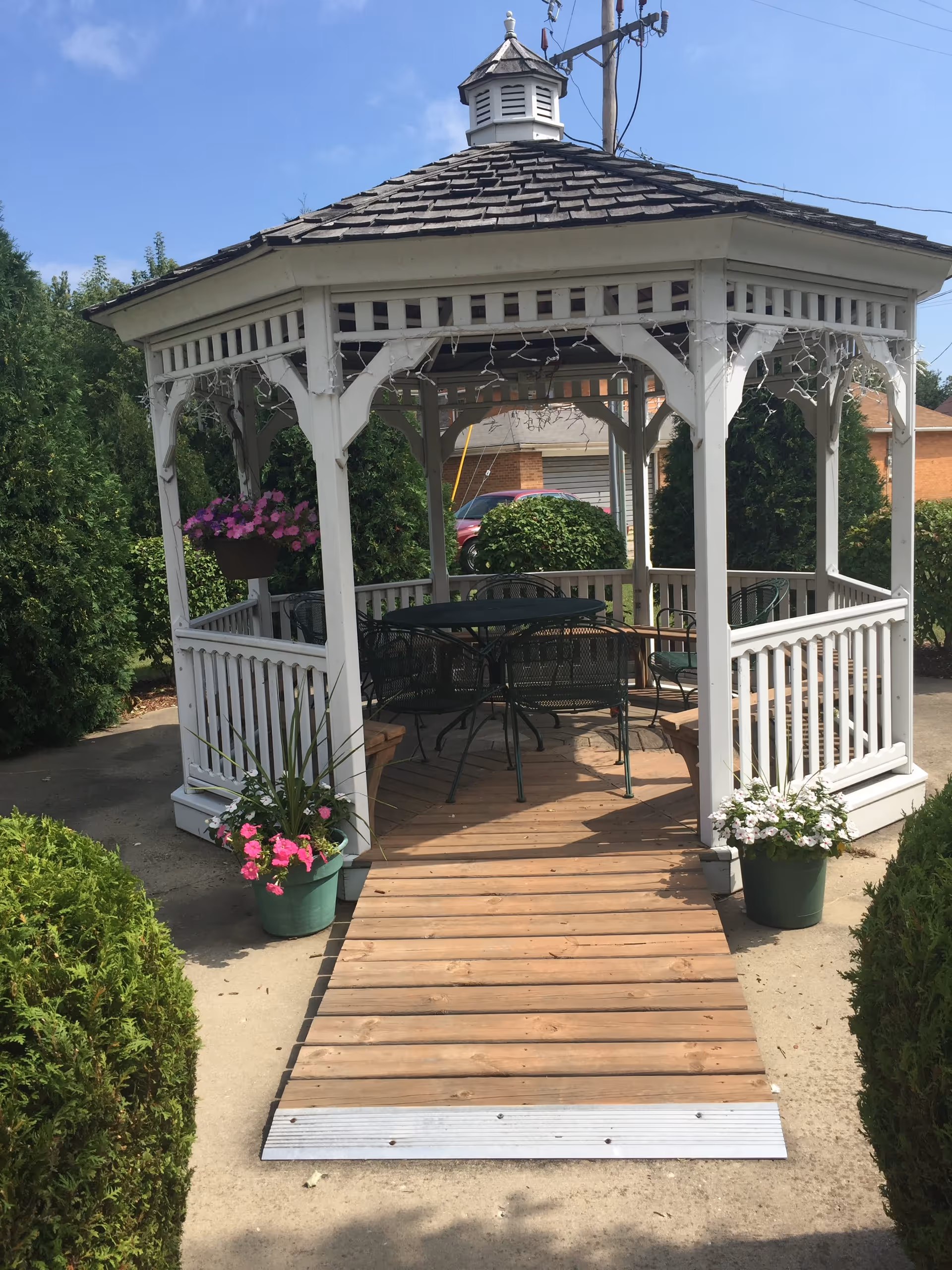 A white wooden gazebo with a shingled roof and decorative trim, surrounded by green bushes and potted flowers. Inside the gazebo, there is a metal table with several matching chairs. A wooden ramp leads up to the entrance of the gazebo.