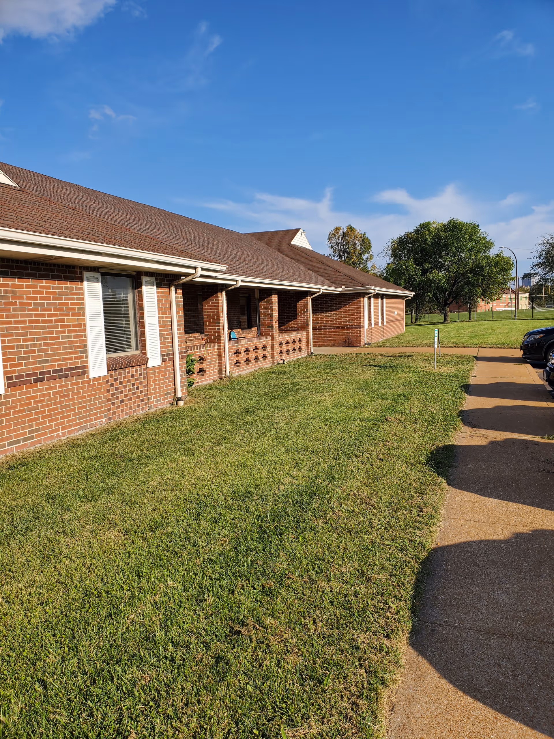 Single-story brick building with a pitched roof, front lawn, sidewalk, and parked cars under a blue sky.