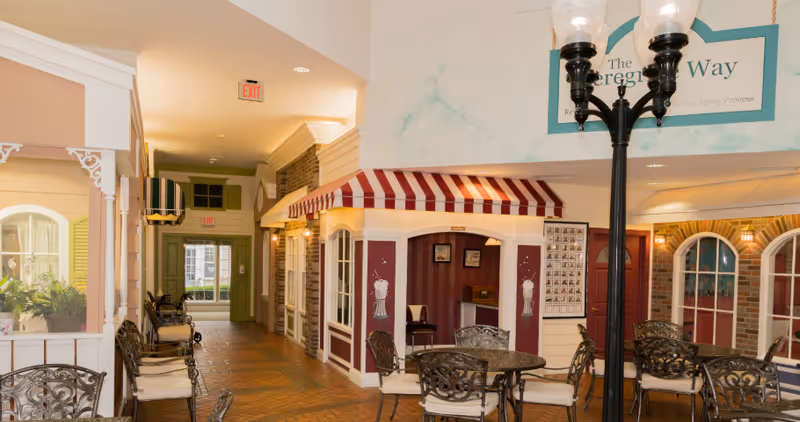 Indoor common area of a senior living facility with café-style storefront facades, round tables and chairs along a hallway.