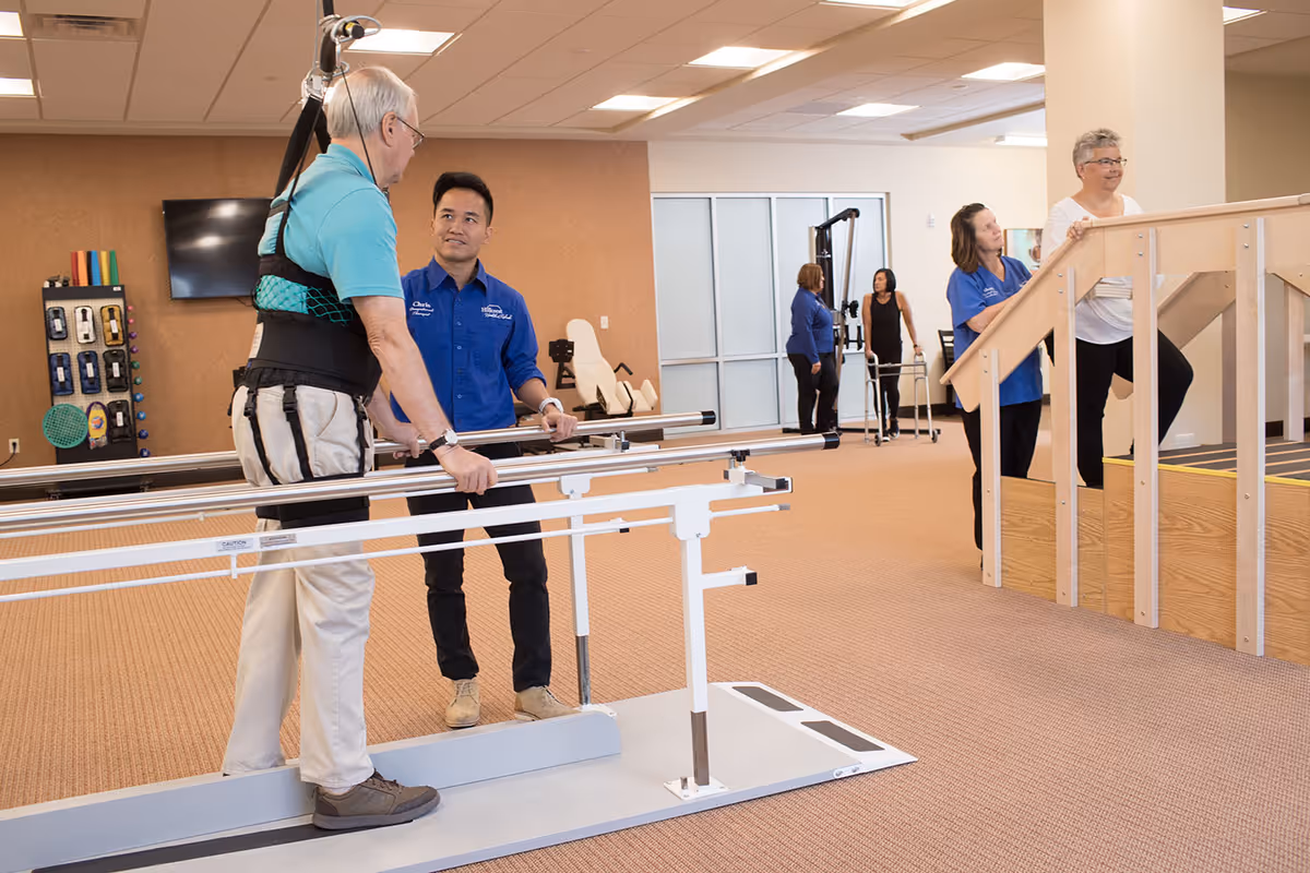 An elderly man wearing a support harness is assisted by a staff member while walking between parallel bars in a rehabilitation or physical therapy room. In the background, another elderly woman is walking up wooden stairs with the help of a staff member. The room has beige carpeting and various therapy equipment.