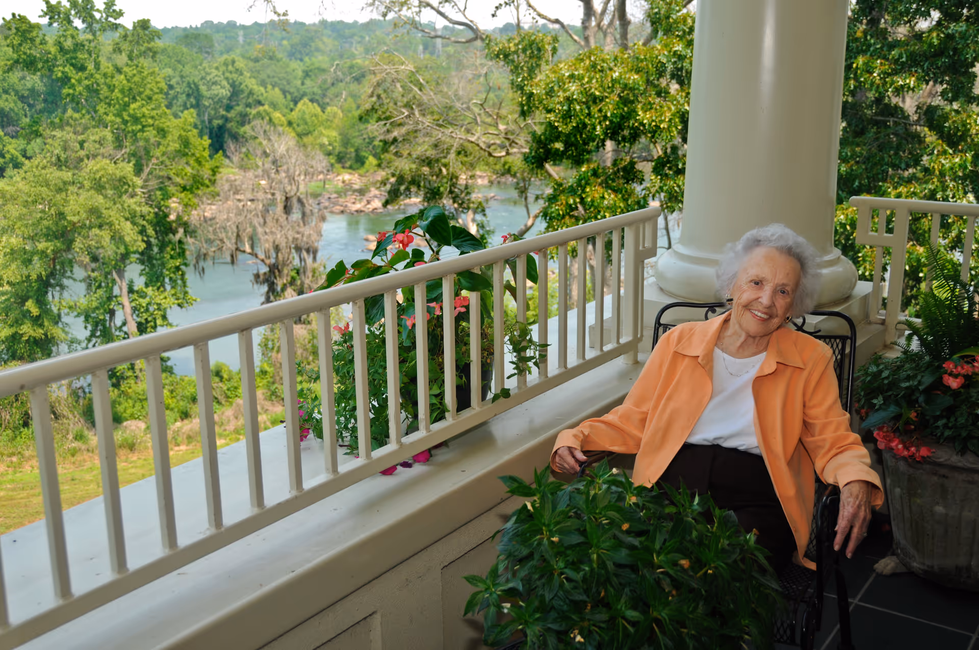 An elderly woman in an orange jacket sits on a balcony surrounded by plants with a scenic view of trees and water in the background.