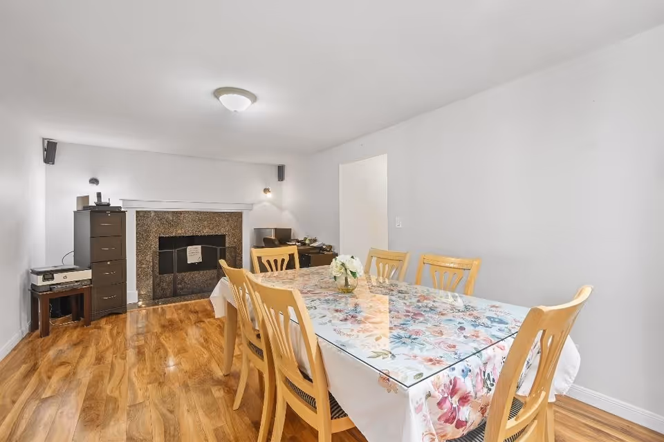 A dining room with a wooden table covered by a floral tablecloth and a glass top, surrounded by six wooden chairs. The room has light-colored walls, a ceiling light fixture, and a fireplace with a granite surround. There is a tall dark brown cabinet and a small table with a printer on the left side of the fireplace, and a desk with a chair in the corner.