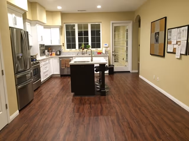A modern kitchen with wooden flooring, white cabinets, stainless steel refrigerator and oven, a central island with a sink, and two black bar stools. There is a window above the sink and a bulletin board on the right wall.