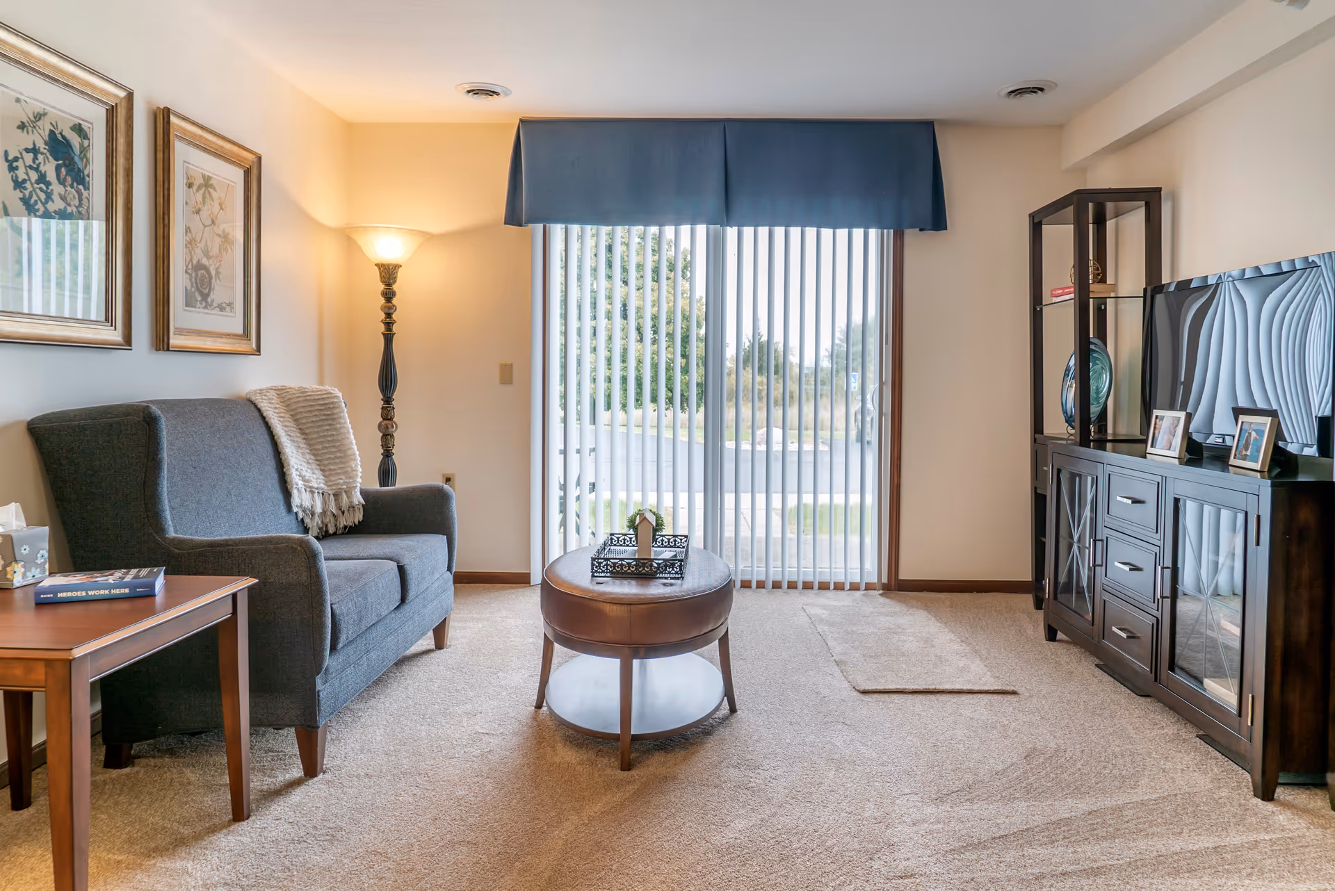 A cozy living room with a gray upholstered loveseat, a wooden side table with a book and tissue box, a round wooden coffee table with a decorative tray, a tall floor lamp, and a dark wooden TV stand with a flat-screen TV and framed photos. Vertical blinds cover a sliding glass door that opens to an outdoor view.