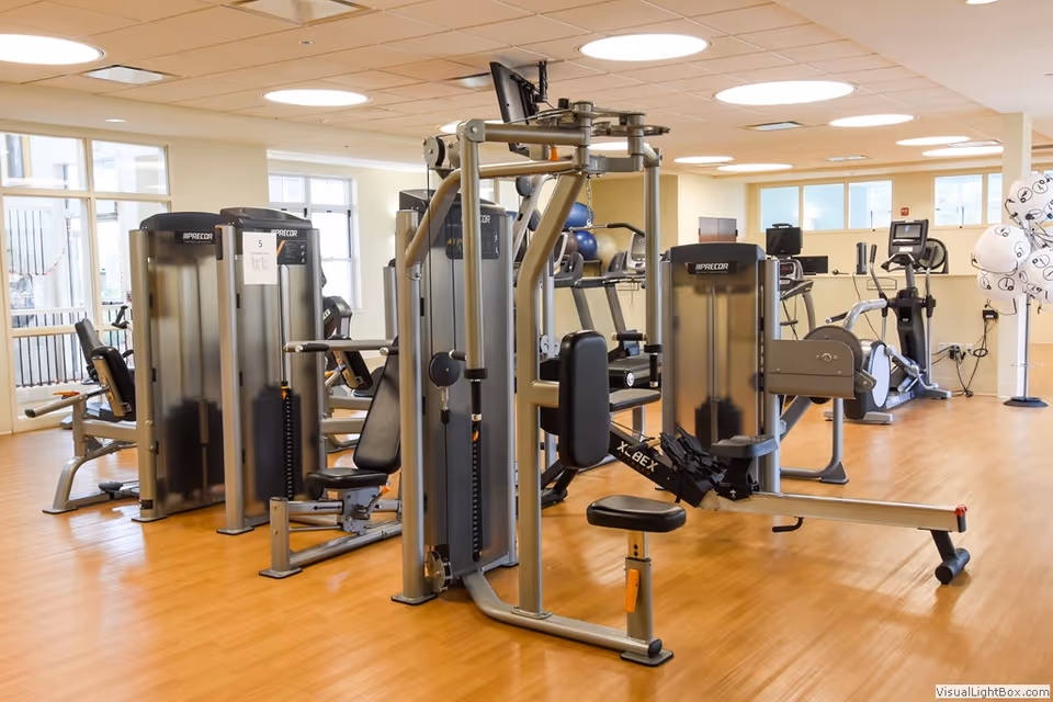 A bright and spacious fitness room with various exercise machines including weight machines, a rowing machine, and cardio equipment. The room has large windows allowing natural light to fill the space and a light wood floor.