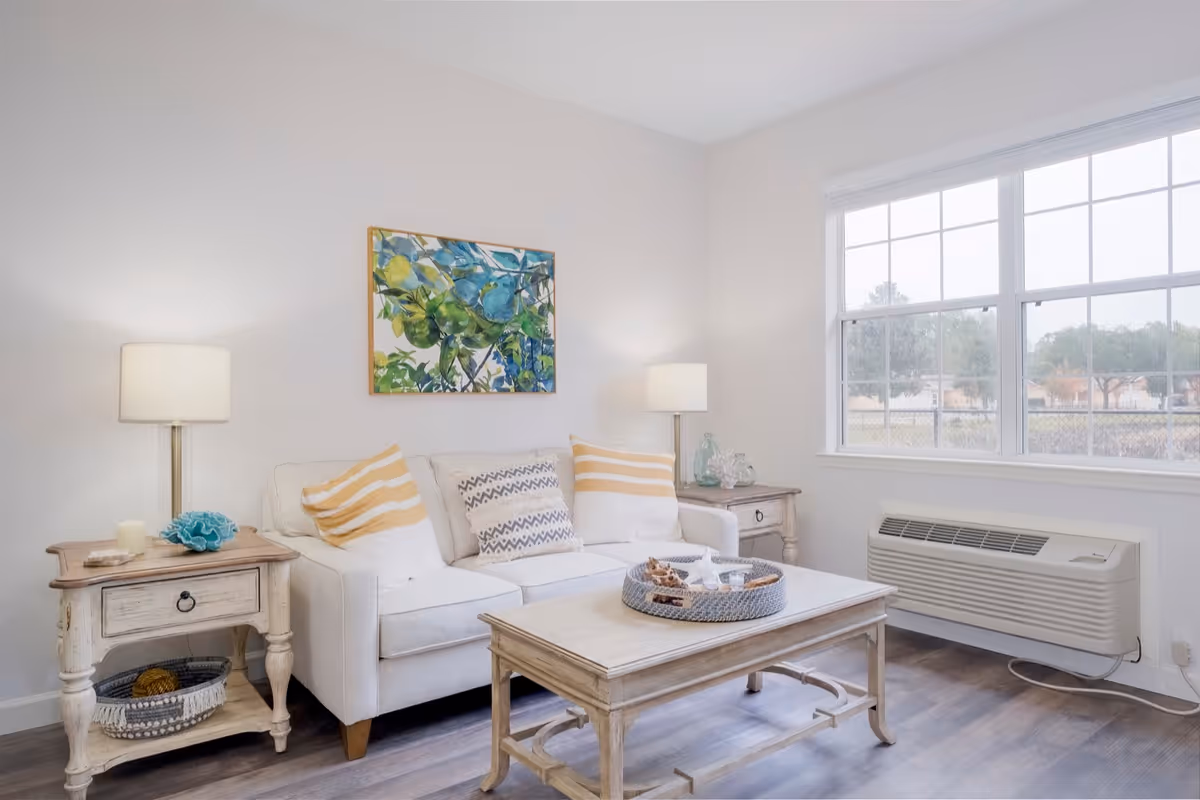 A bright and cozy living room with a white sofa adorned with striped and patterned pillows, two wooden side tables each with a lamp, a wooden coffee table with a decorative tray, a large window letting in natural light, and a colorful painting of green and blue leaves on the wall.