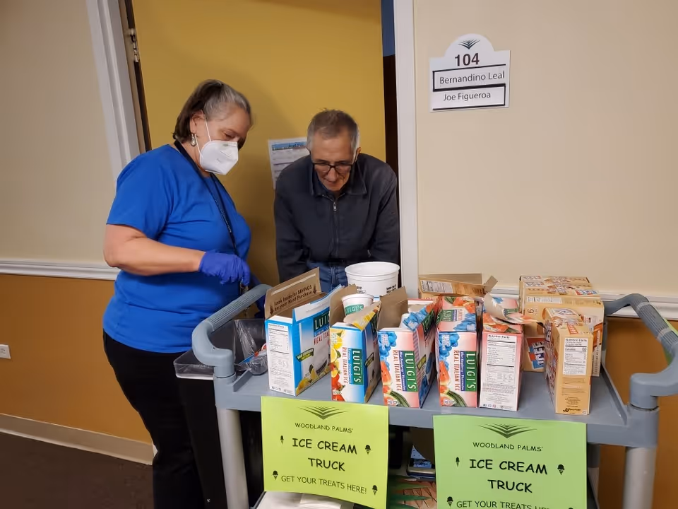 A staff member wearing a blue shirt, gloves, and a face mask is assisting an elderly man standing behind a cart filled with various ice cream products. The cart has two green signs that read 'Woodland Palms Ice Cream Truck Get Your Treats Here!'. They are in a hallway outside a room labeled 104 with the names Bernandino Leal and Joe Figueroa on the door sign.