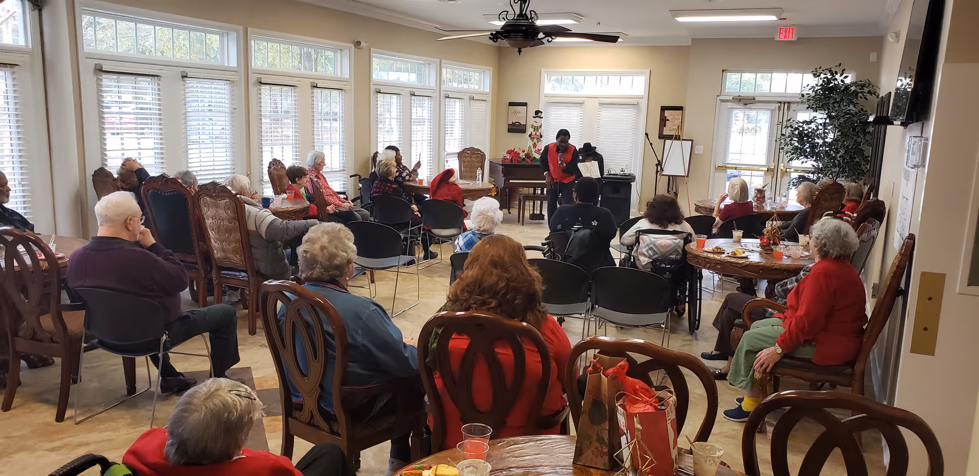 A group of elderly residents seated around tables and chairs in a bright common room watching a presenter at the front.