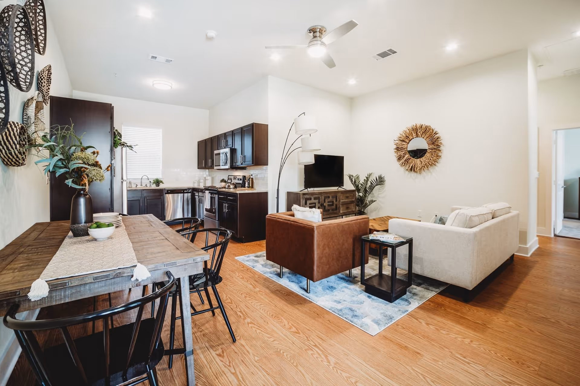 A bright and modern open-concept living space featuring a kitchen with dark wood cabinets, stainless steel appliances, and a dining area with a wooden table and black chairs. The living area includes a beige sofa, a brown armchair, a TV on a wooden stand, a floor lamp, and decorative wall art with a round mirror. The room has wooden flooring and white walls with recessed lighting and a ceiling fan.