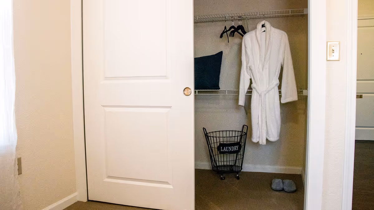 A partially open white closet door reveals a closet interior with a white bathrobe hanging on a hanger, a black laundry basket labeled 'LAUNDRY', a dark pillow on the shelf, several empty black hangers, and a pair of gray slippers on the floor.