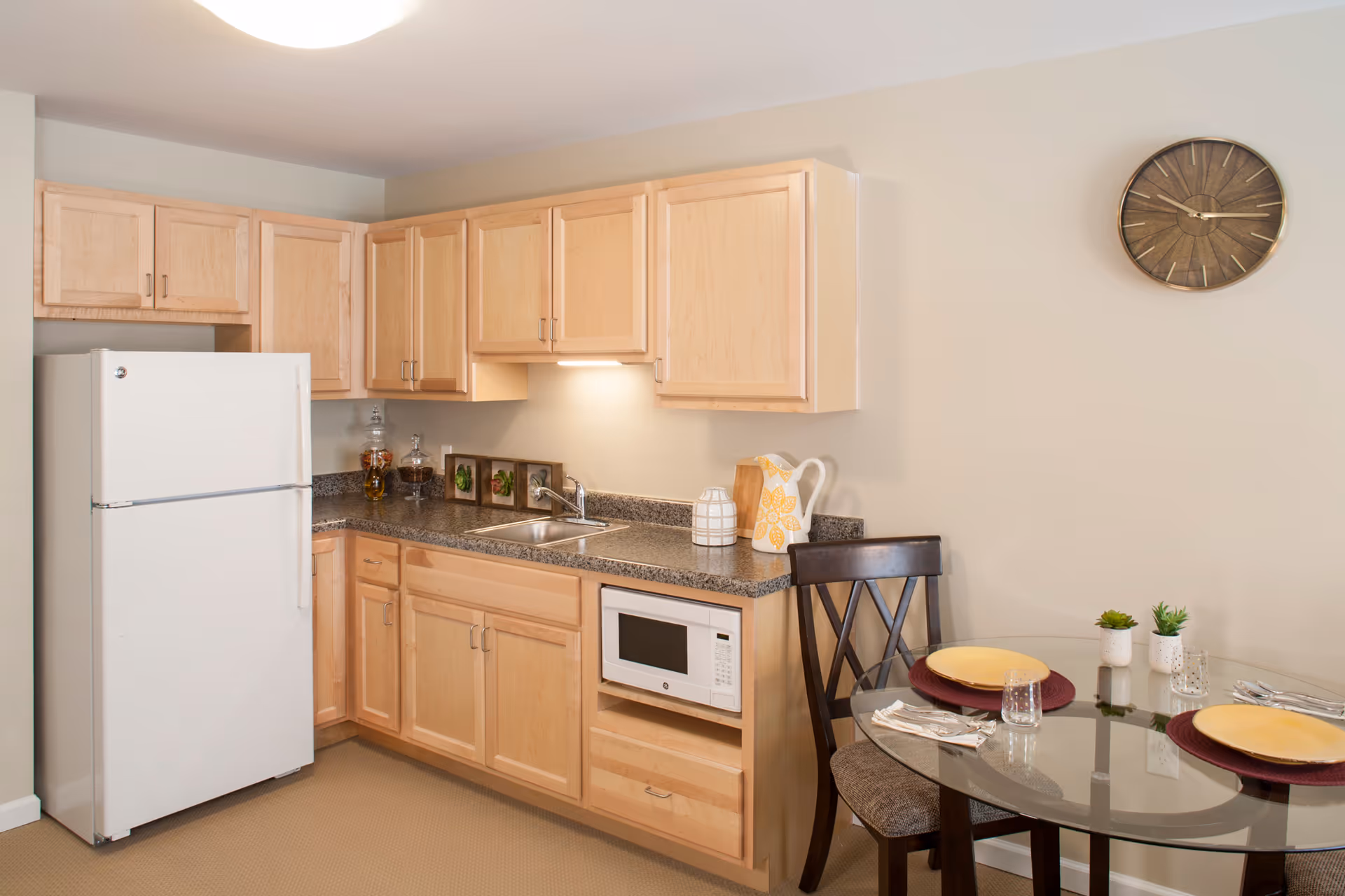 A small kitchen area with light wood cabinets, a white refrigerator, a microwave, and a granite countertop with a sink. To the right, there is a glass-top dining table set for two with yellow plates, glasses, and silverware. A wooden chair is placed at the table, and a round wall clock hangs on the beige wall above.