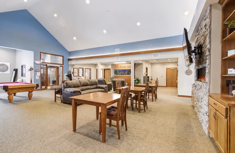 A spacious common area in a senior living facility featuring a stone fireplace with a mounted TV, wooden tables and chairs, a gray couch, a pool table, and a high vaulted ceiling with recessed lighting. The walls are painted in neutral tones with a blue accent wall, and there are large windows and doors in the background.