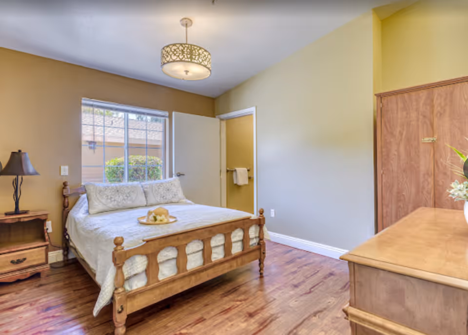 A cozy bedroom with a wooden bed frame and white bedding, a wooden nightstand with a lamp, a window showing greenery outside, a wooden dresser with a vase of flowers, and a door leading to another room with a towel hanging on a rack.