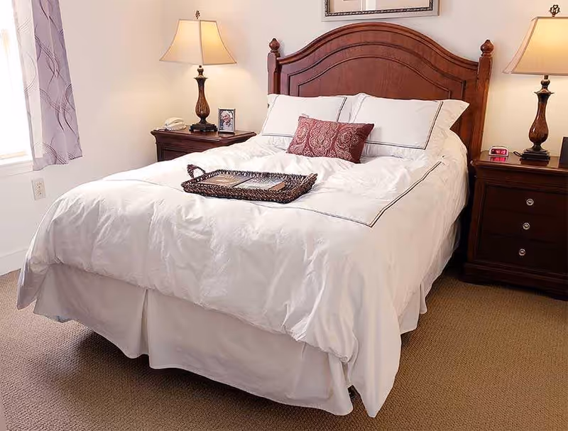 A neatly made bedroom with a white bed, wooden headboard, matching bedside tables and lamps.