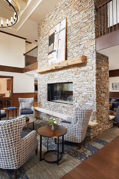 A cozy senior living facility lounge area featuring a large stone fireplace with a wooden mantel and abstract artwork above it. The seating area includes patterned armchairs arranged around small round tables with a vase of flowers on one table. The space has high ceilings with exposed beams and a chandelier, creating a warm and inviting atmosphere.