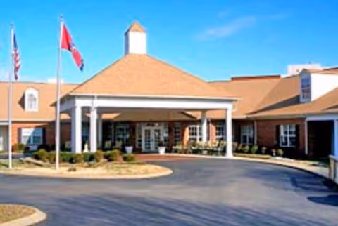 Brick senior living facility exterior with a covered entrance and flagpoles by a circular driveway.