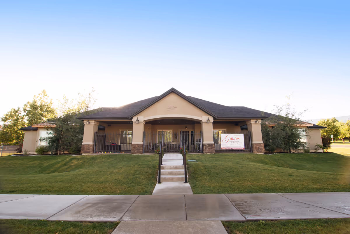 Front exterior view of The Gables Assisted Living of North Logan building with a walkway leading up to the entrance, surrounded by green grass and trees under a clear blue sky.