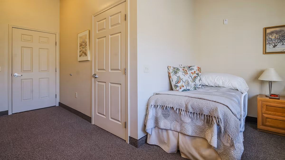 A small, neatly made single bed with a floral pillow and a light gray blanket in a room with beige walls and carpeted floor. Next to the bed is a wooden nightstand with a lamp and a digital clock. Two closed doors and a framed picture are visible on the opposite wall.