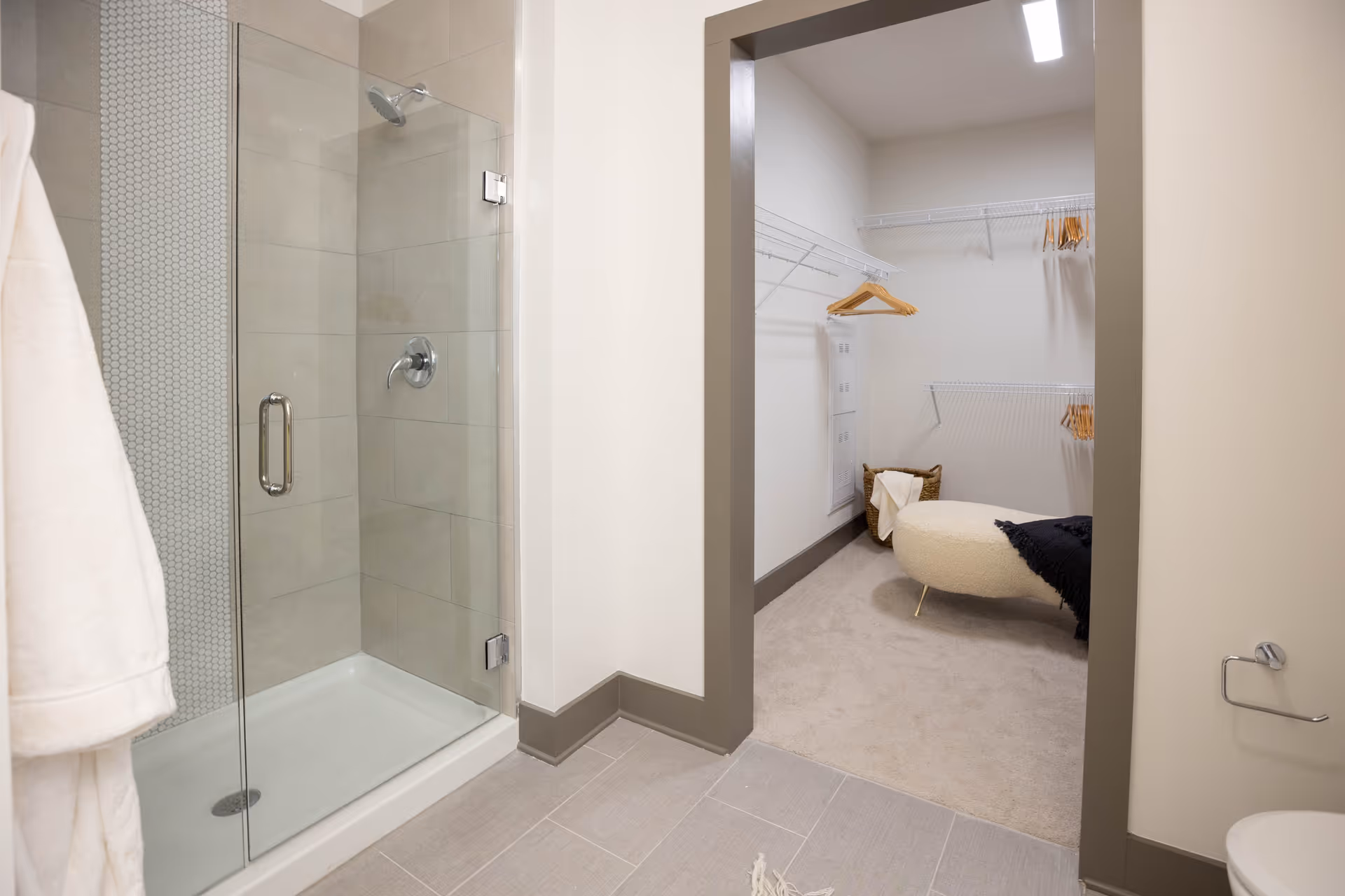 A modern bathroom with a glass-enclosed shower and an open doorway into a walk-in closet with shelving and a round ottoman.