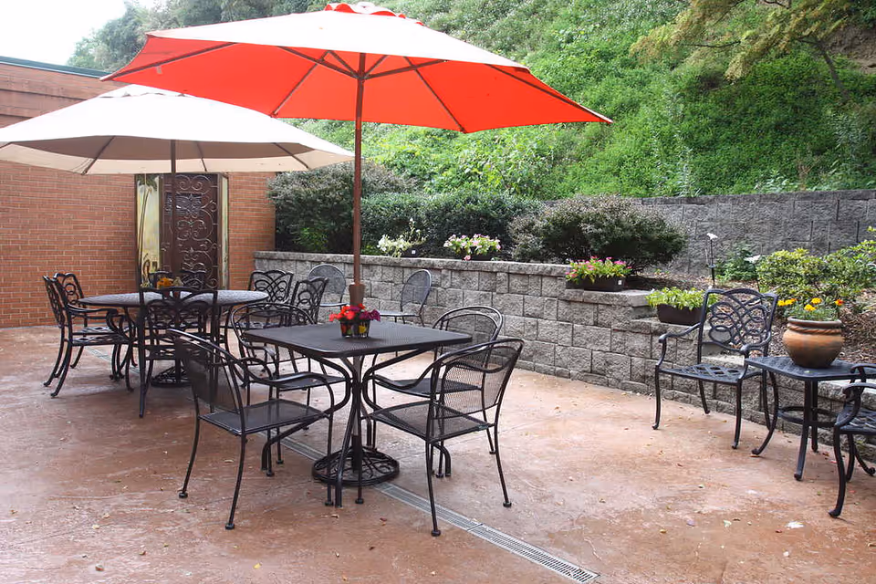 Outdoor patio area with metal tables and chairs, two large umbrellas providing shade, potted plants on tables and along a stone retaining wall, surrounded by greenery.