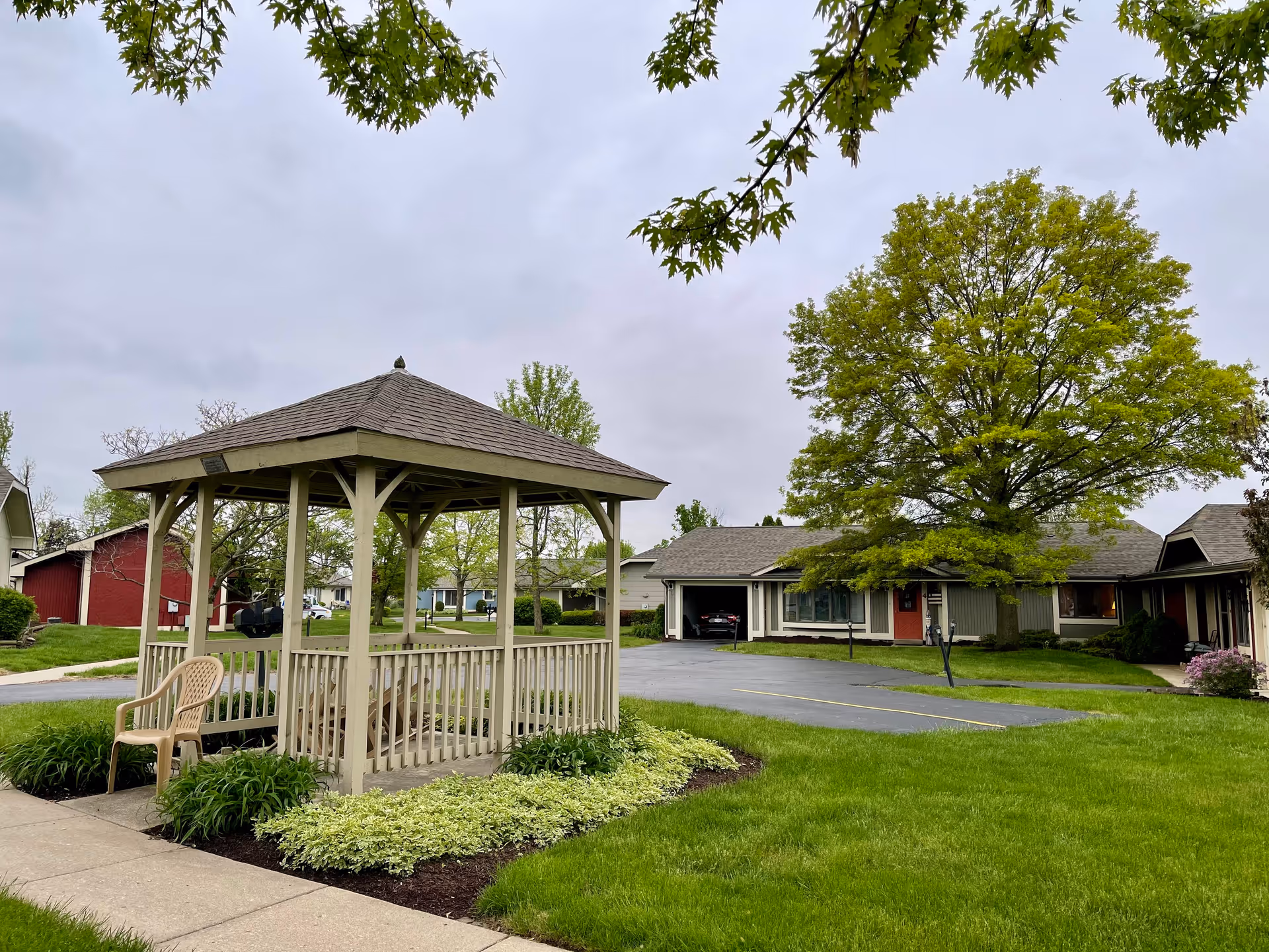 A wooden gazebo on a landscaped lawn in front of single-story cottages and a large tree under an overcast sky.