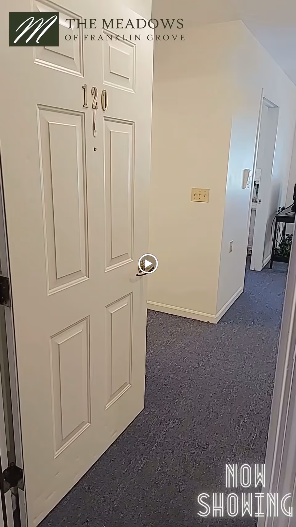 Open white door numbered 120 leading into a hallway with blue carpet and white walls inside The Meadows of Franklin Grove Assisted Living facility.