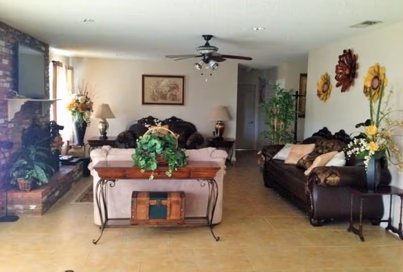 A cozy living room with two dark leather sofas and one light-colored sofa arranged around a wooden table with a green plant. The room features a brick fireplace with plants on the hearth, a ceiling fan with lights, framed artwork on the wall, table lamps, and decorative flower wall hangings. The floor is tiled and there are additional plants and side tables around the room.