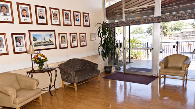 A reception lobby with chairs and a small table, framed portraits on the wall, a potted plant, and glass entrance doors.