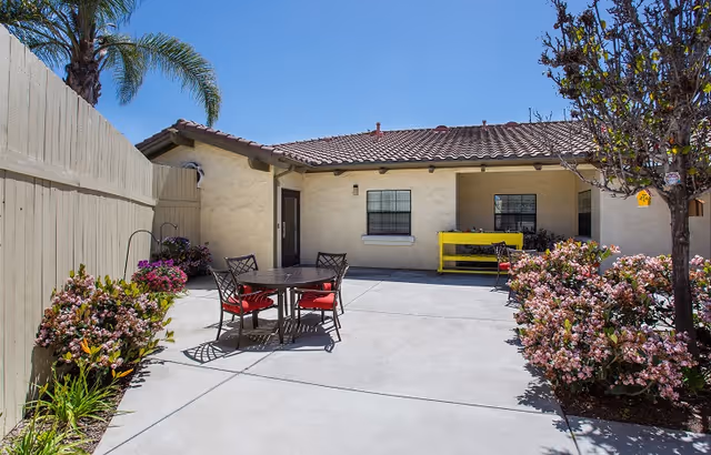 Outdoor patio area at Point Loma Estates Memory Care with a round table and four chairs with red cushions, surrounded by flowering bushes and a tree. The building has a beige stucco exterior with a tiled roof and two windows. A bright yellow bench is placed against the wall under one window.