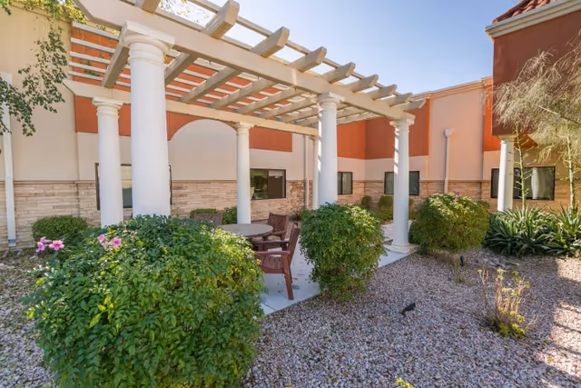 Outdoor seating area with a pergola supported by white columns, surrounded by bushes and gravel landscaping, adjacent to a building with beige and orange walls and several windows.