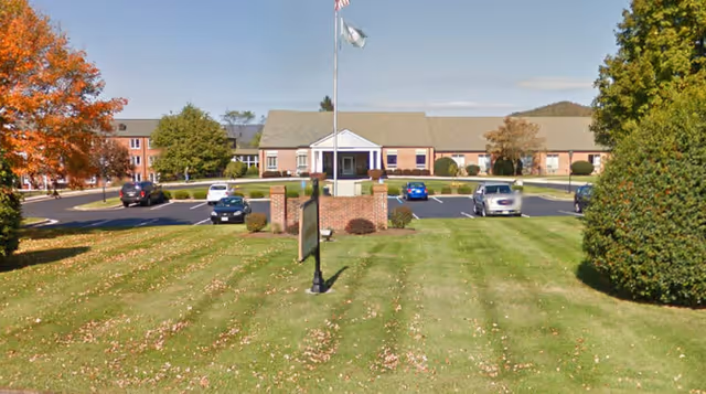 Front exterior view of Carrington Place at Wytheville, a single-story brick building with a central entrance featuring white columns. The building is surrounded by a parking lot with several cars and a well-maintained lawn with trees showing autumn foliage.