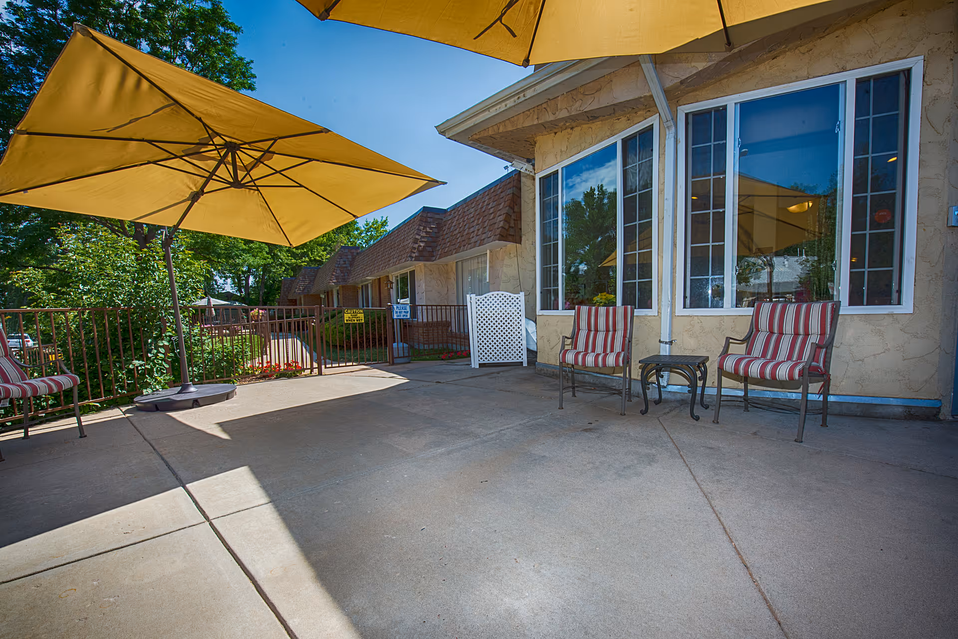Outdoor patio area at Clear Creek Care Center with two large yellow umbrellas providing shade over a concrete floor. There are two striped cushioned chairs and a small glass-top table near a building with large windows. Trees and greenery are visible in the background.
