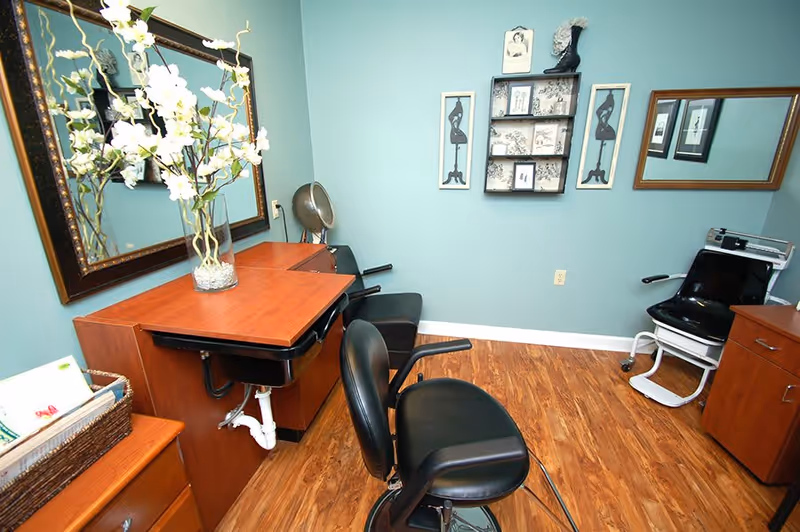 Interior of a salon or grooming room with two black salon chairs, a wooden counter with a large mirror, a vase with white flowers, and framed artwork on light blue walls. The floor is wood laminate.