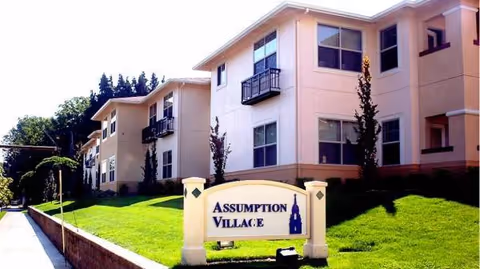 Exterior view of Assumption Village with sign and green lawn in front.