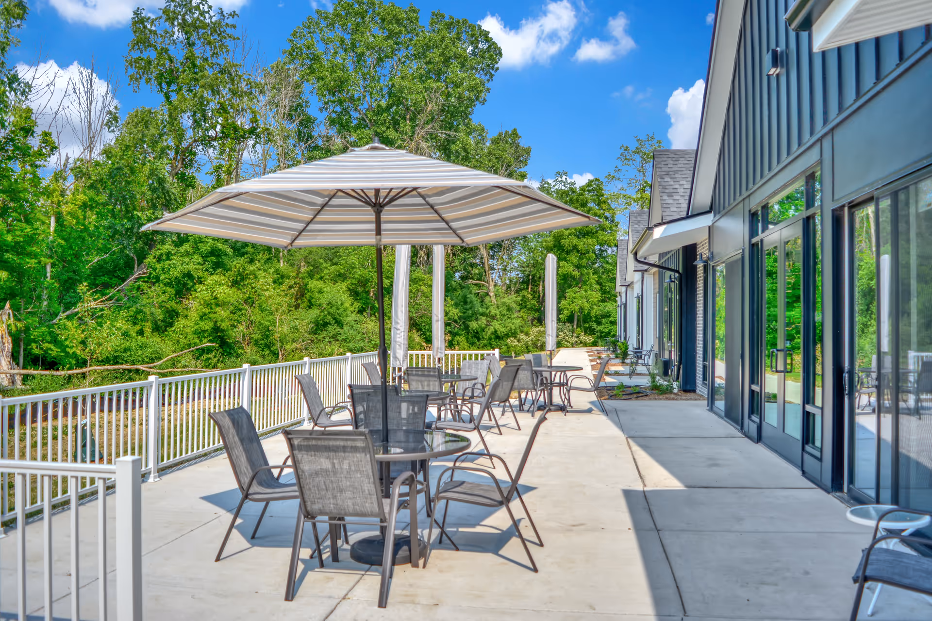 Outdoor patio area with several tables and chairs, each table shaded by large umbrellas. The patio is adjacent to a building with large glass doors and windows, surrounded by lush green trees under a clear blue sky.