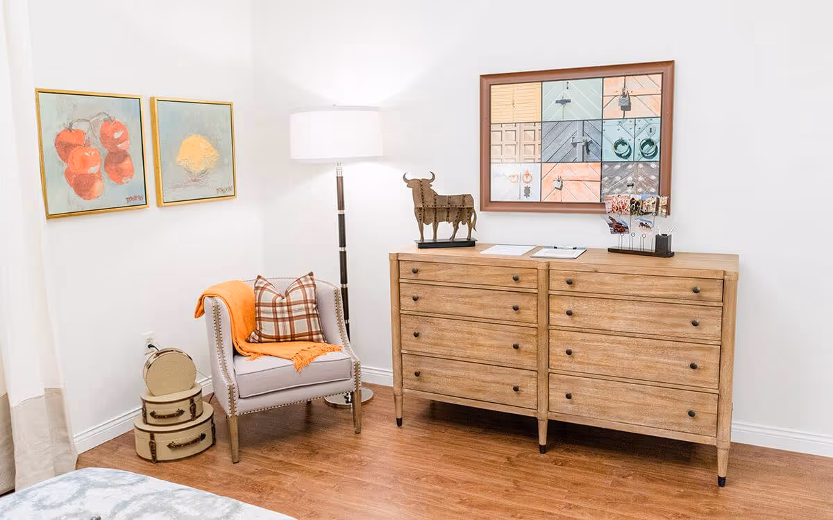 A cozy corner of a bedroom featuring a wooden dresser with multiple drawers, a decorative metal bull sculpture, and a display of postcards. Next to the dresser is a cushioned armchair with a plaid pillow and an orange throw blanket. The walls are adorned with two framed paintings of fruit and a larger framed artwork above the dresser. The floor is wooden, and a tall floor lamp provides soft lighting.