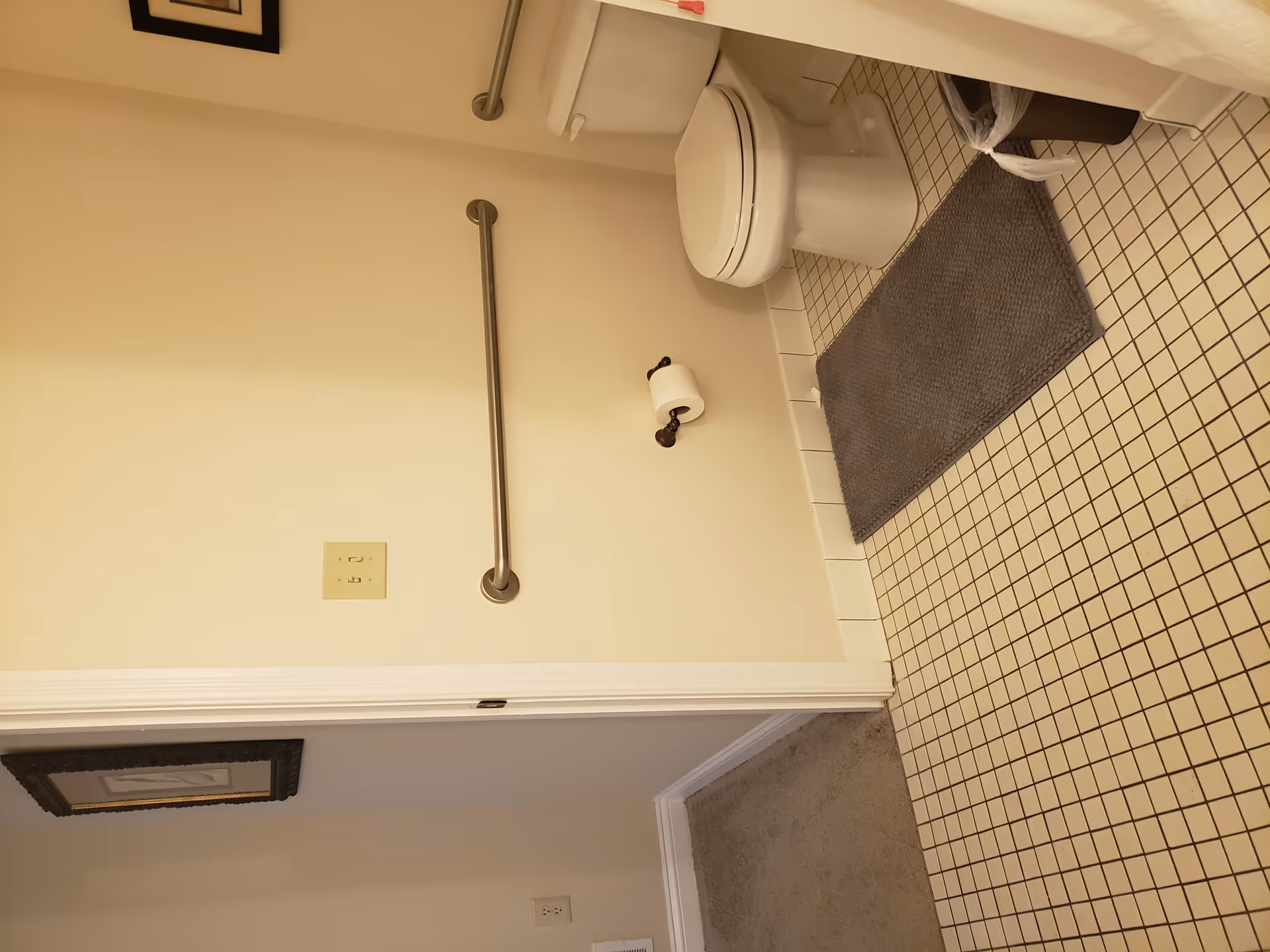 Bathroom corner showing a toilet, wall-mounted grab bar, tiled floor and a gray bath mat.