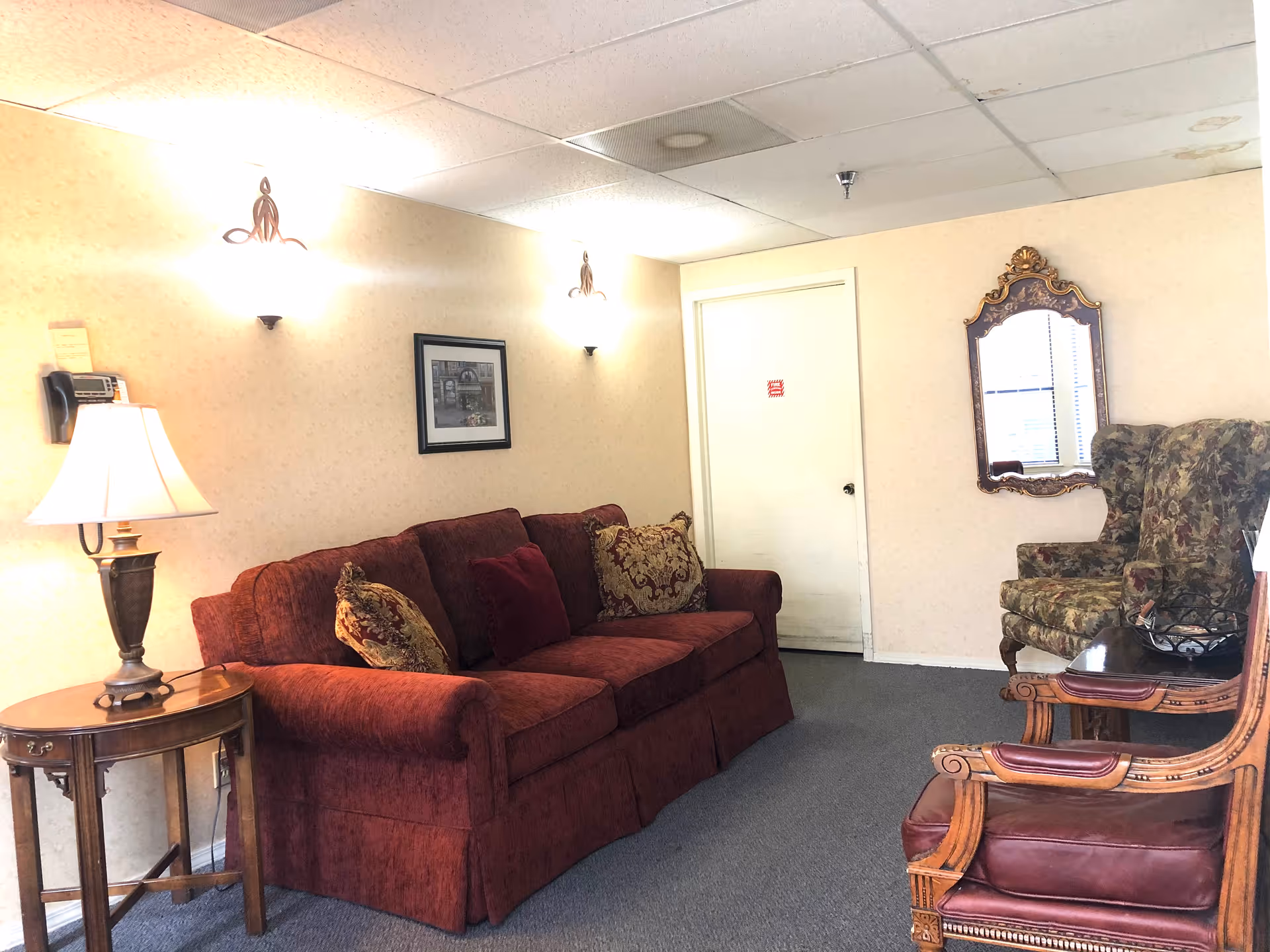 A cozy sitting area in a retirement community featuring a red upholstered sofa with decorative pillows, a wooden side table with a lamp, two patterned armchairs, a wooden coffee table, a wall-mounted mirror with an ornate frame, and a framed picture on the wall. The room has beige walls, a carpeted floor, and ceiling tiles with some stains.