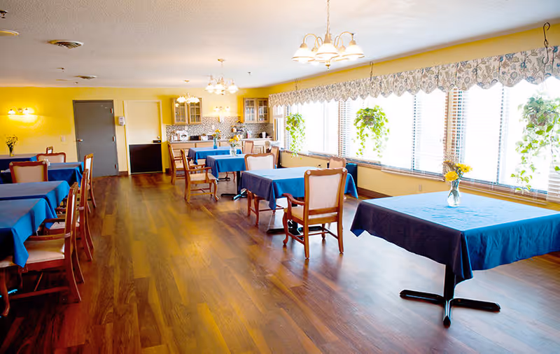 A bright dining room with wooden floors, several tables covered with blue tablecloths, and wooden chairs. Large windows on one side let in natural light and have hanging plants and floral valances. The back wall features a kitchenette area with cabinets, a microwave, and a coffee maker.