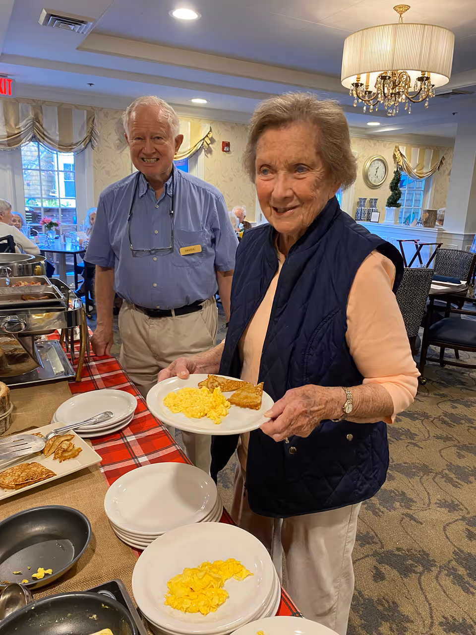 An elderly woman holding a plate with scrambled eggs and a piece of toast stands at a buffet table with food trays and plates. An elderly man wearing a name tag stands behind her, smiling. The setting is a dining room with tables, chairs, and a chandelier overhead.
