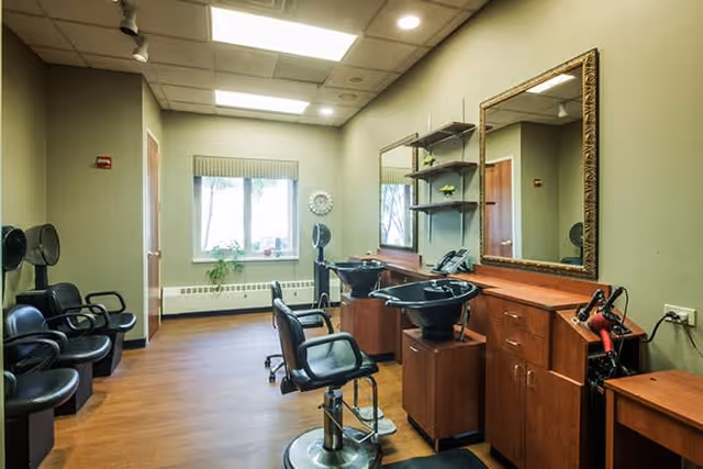Interior view of a hair salon area with multiple black salon chairs, hair washing stations, large mirror, wooden cabinets, and a window with a small plant on the windowsill.