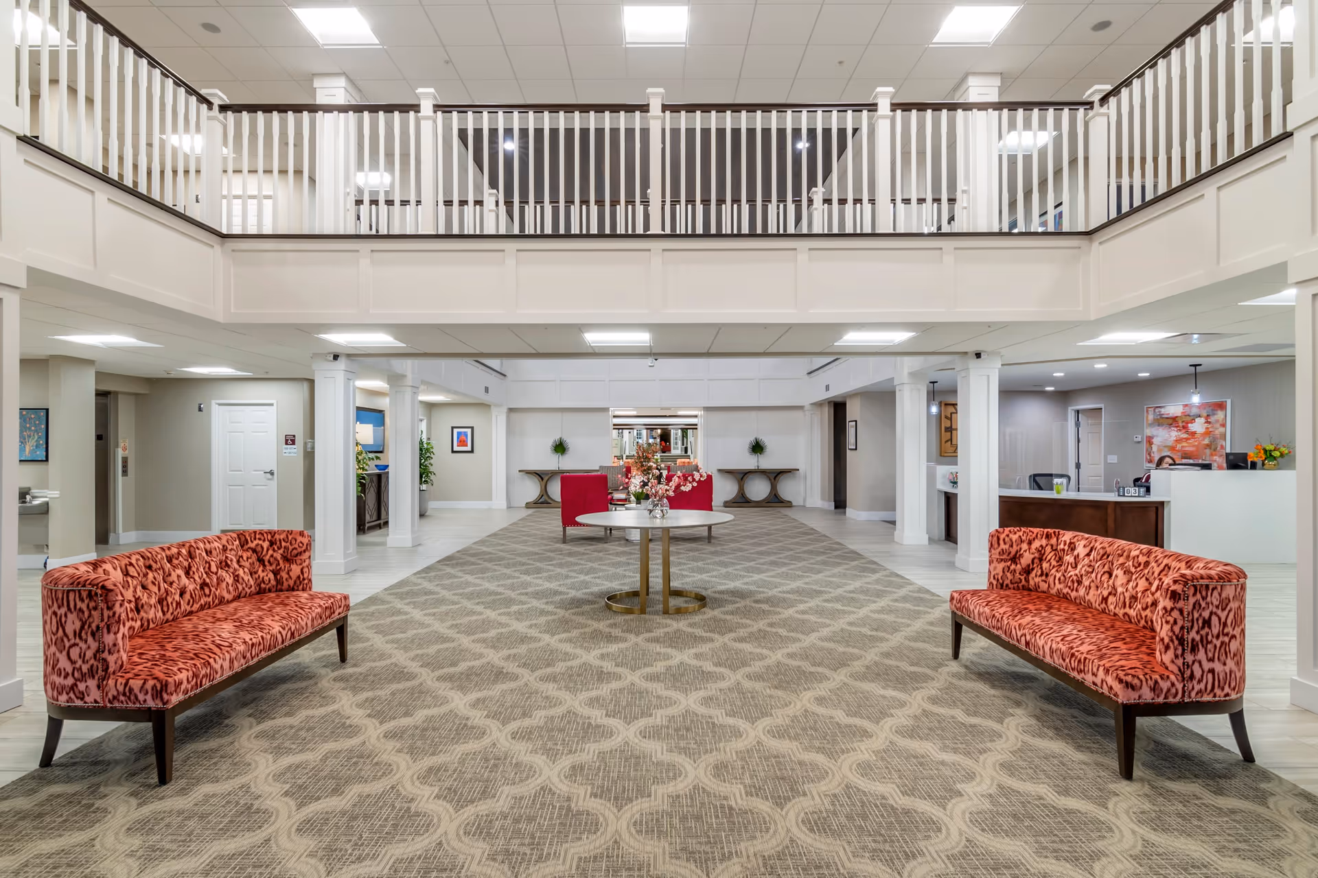 Spacious senior living community lobby with two red patterned sofas facing each other on a large patterned carpet. A round table with a flower arrangement is centered between the sofas. The lobby has white walls, columns, and a second-floor balcony with white railings overlooking the area. There is a reception desk on the right side and decorative tables with plants along the back wall.