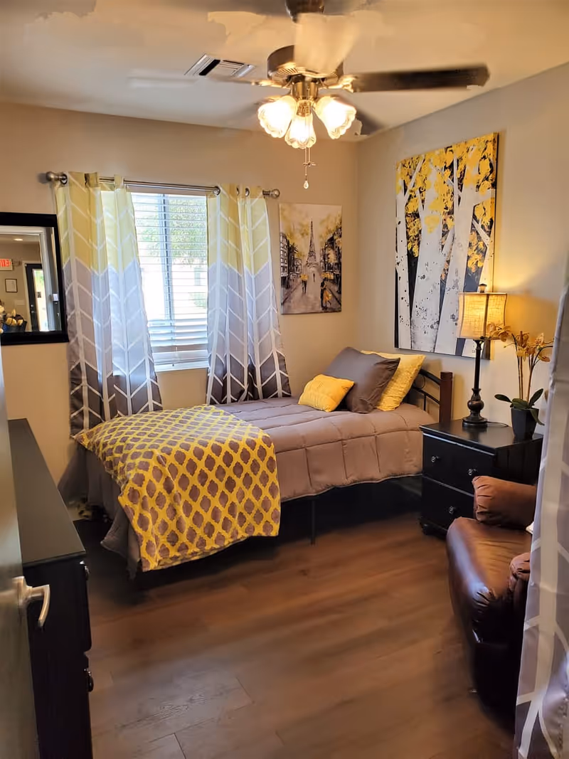 A cozy bedroom with a single bed covered in a gray comforter and yellow patterned blanket. The bed has gray and yellow pillows. There is a window with gray and yellow curtains, a ceiling fan with lights, a black nightstand with a lamp and a small plant, a brown leather armchair, and two paintings on the walls, one depicting trees with yellow leaves and the other a cityscape.