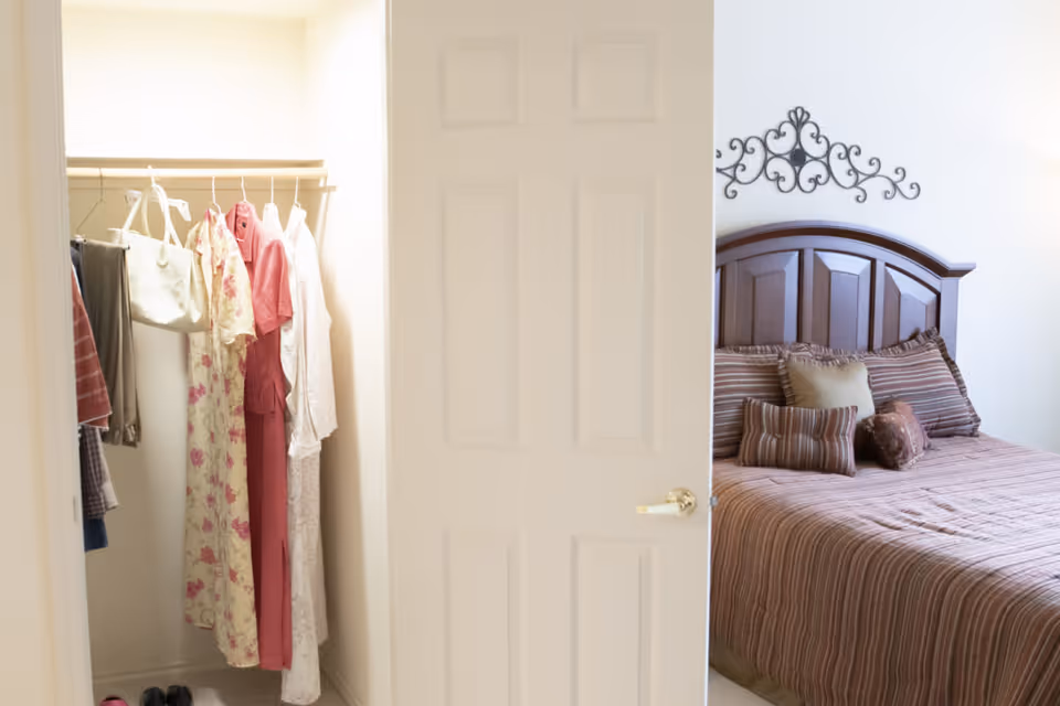 A bedroom with a wooden headboard bed dressed in striped brown bedding and multiple pillows. To the left, an open closet with hanging clothes including dresses and a handbag is visible. The room is softly lit with neutral walls and a decorative metal wall art above the bed.
