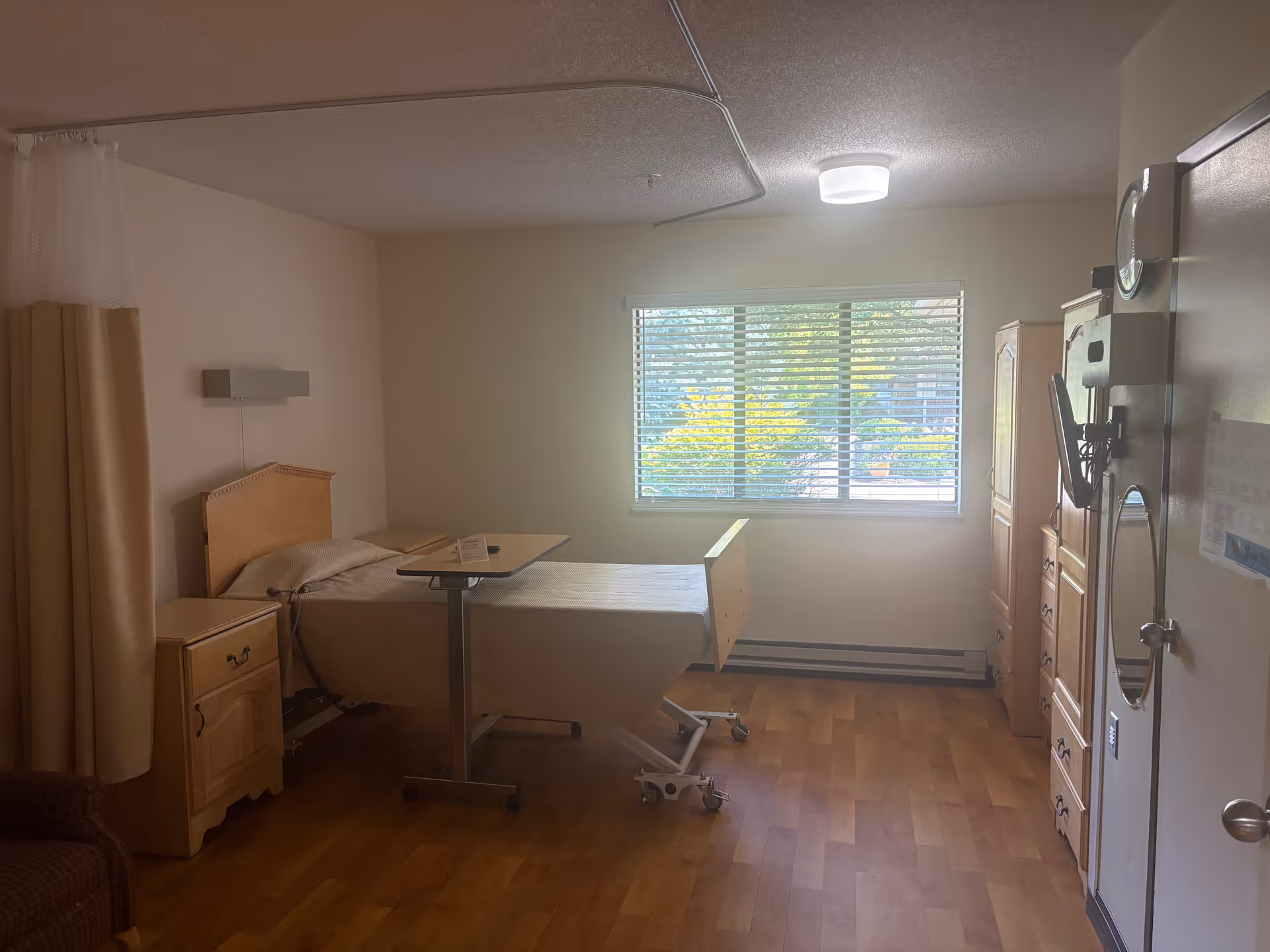 Furnished senior living bedroom with an adjustable hospital-style bed and overbed table, nightstand, dresser, window with blinds, and a privacy curtain.