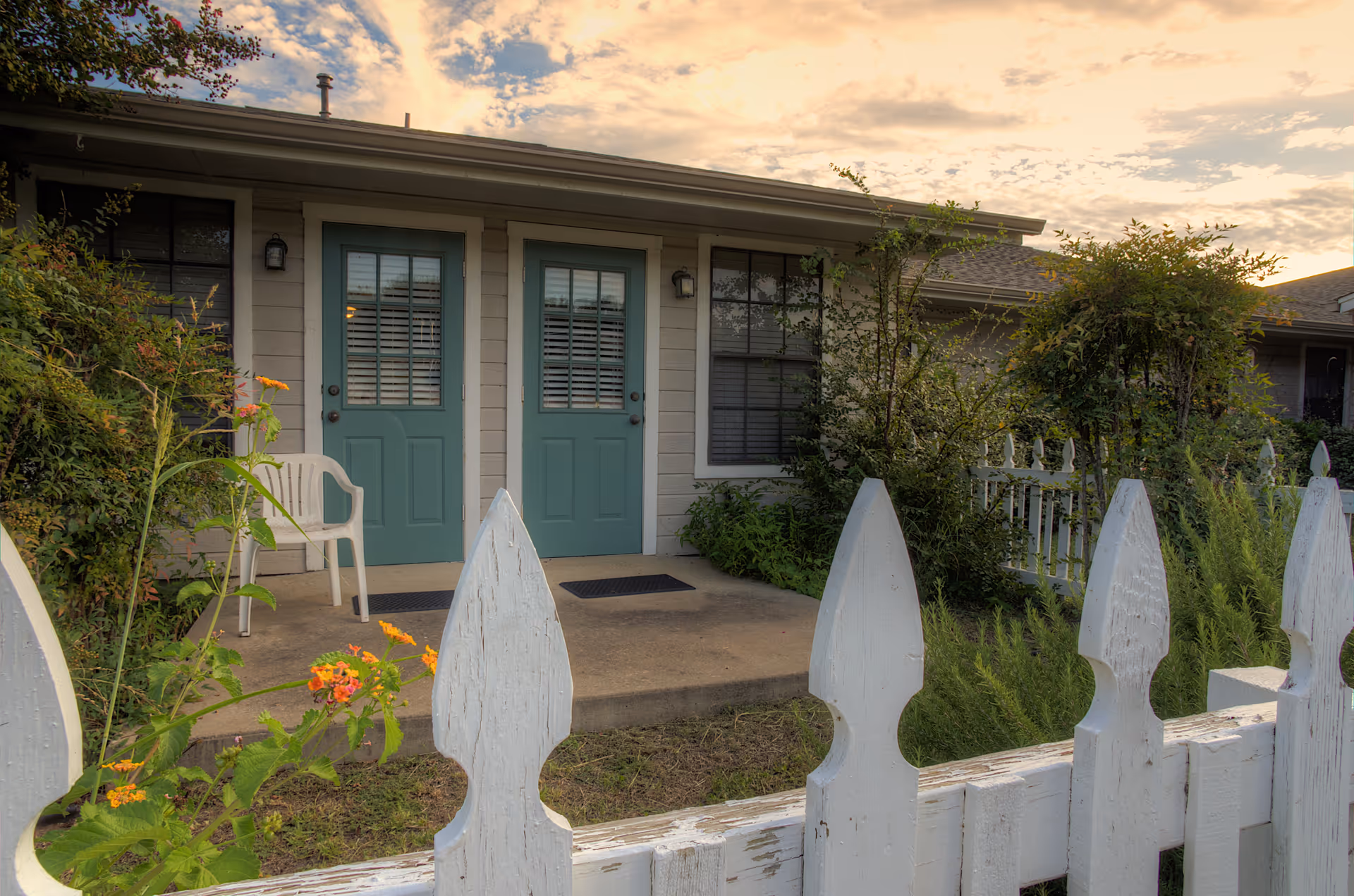 Exterior view of a residential building with two teal-colored doors side by side, each with a window featuring white blinds. A white plastic chair is placed on the concrete porch. The area is surrounded by greenery and a white picket fence in the foreground. The sky is partly cloudy with a warm light from the setting or rising sun.